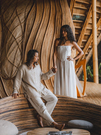 Two people sitting on a large wooden sculpture with a natural setting wearing white Tiyara clothes