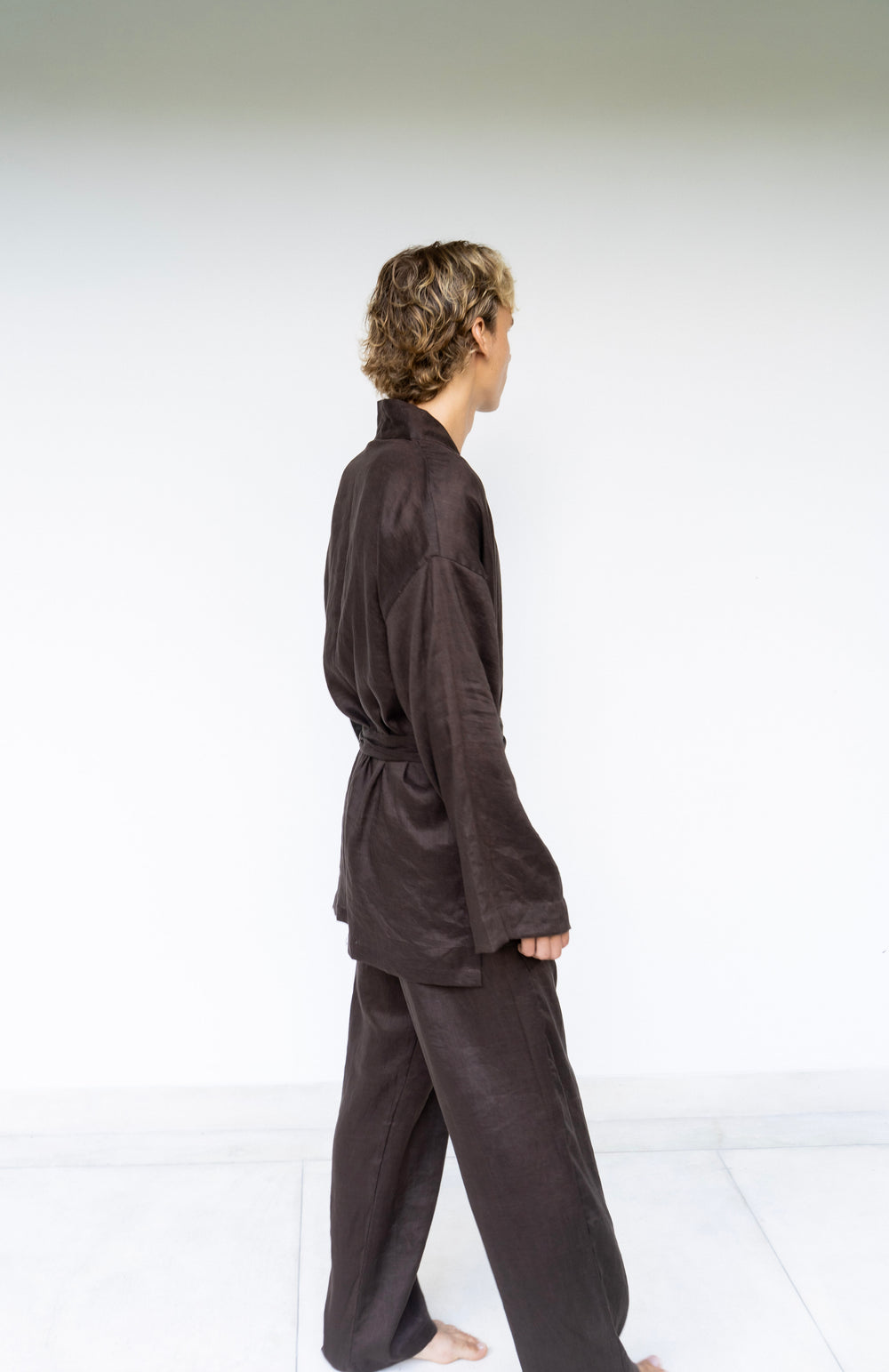 A person wearing a dark brown loose-fitting robe or kimono-style outfit with matching pants, shown from the side against a plain white studio background.