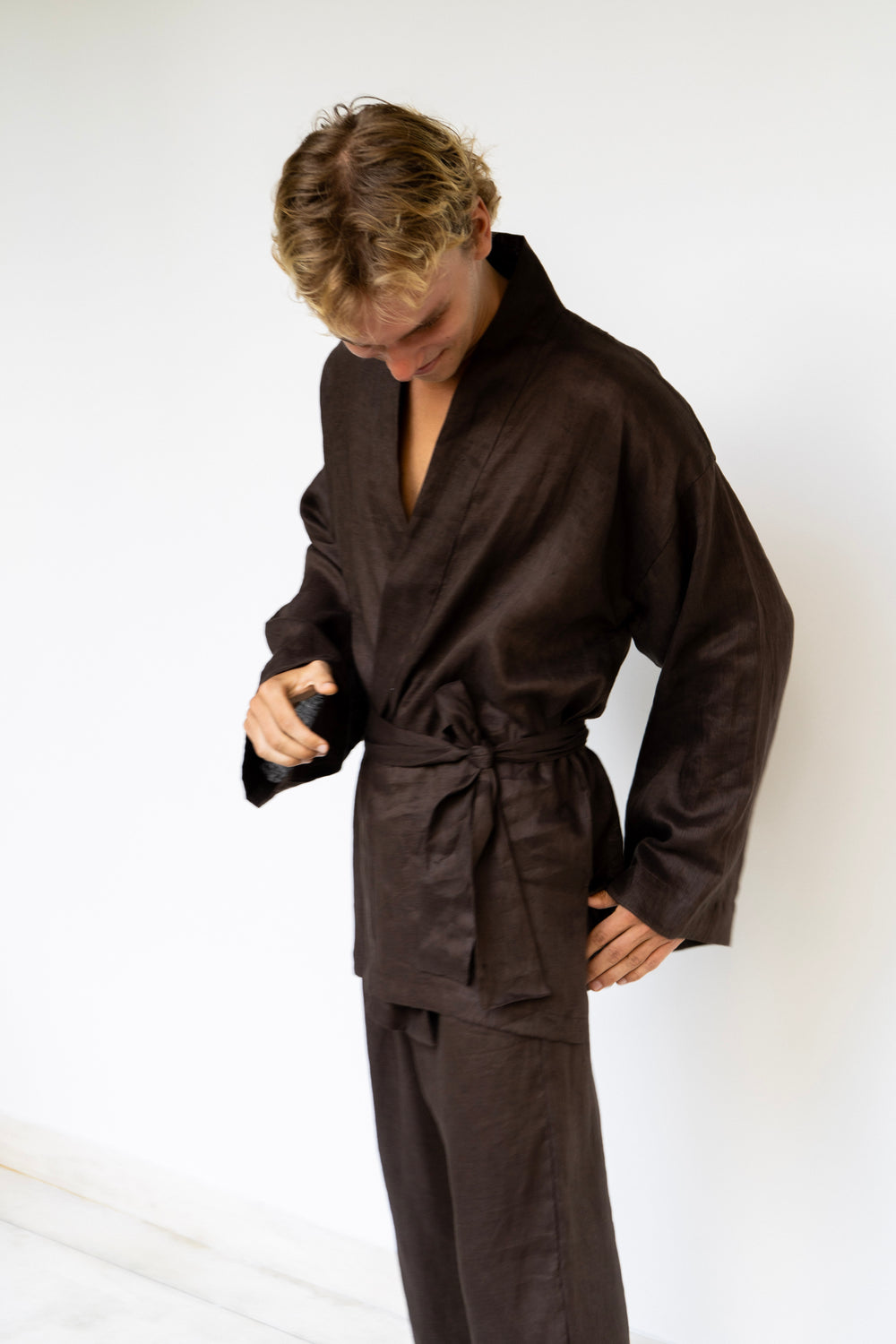A man wearing a Shen kimono & Shen pants in linen dark brown,  looking down while adjusting the collar of the kimono against a plain white background.