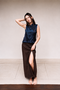 A woman posing in a dark navy blue sleeveless top with ruffles and a dark brown high-slit maxi skirt, standing barefoot against a neutral beige wall.