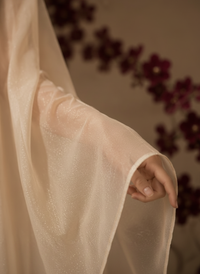 A woman in a flowing, semi-sheer beige gold cape stands in profile against a textured beige wall, with a large dark red vase of branches orchid behind her 