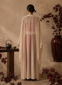 A woman in a flowing, semi-sheer beige gold cape stands in profile against a textured beige wall, with a large dark red vase of branches orchid behind her 