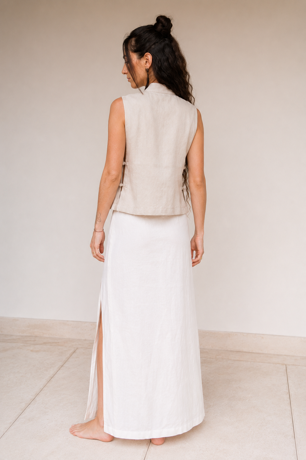 A woman shown from the back wearing a sleeveless light beige vest and a long linen white skirt with a side slit, standing barefoot against a plain light beige wall and floor.